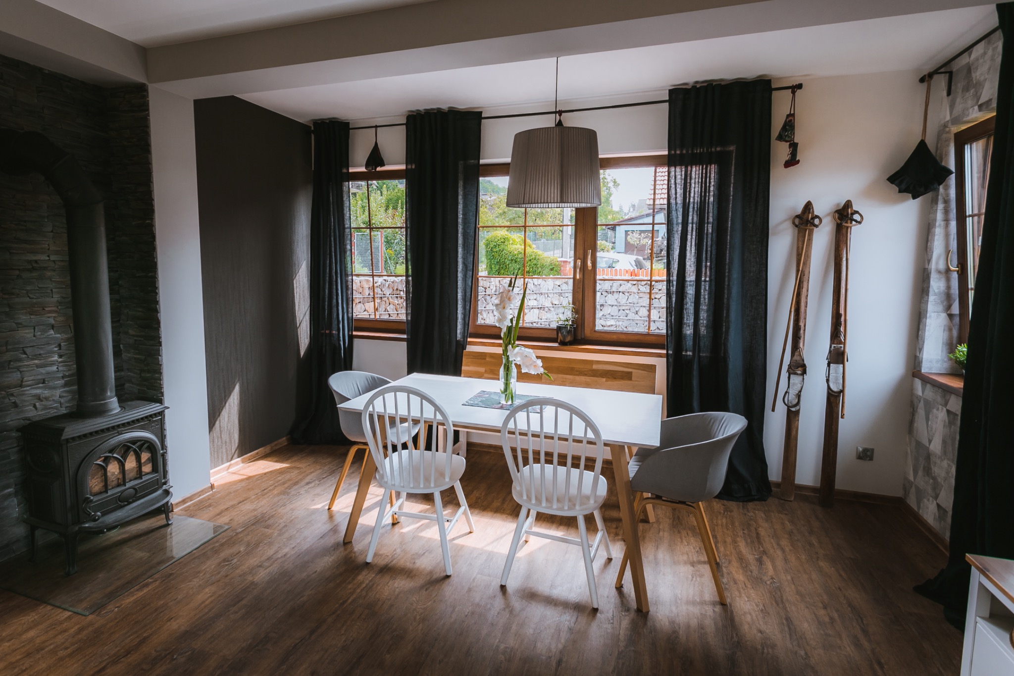 Jasne wnętrze jadalni z białym stołem, krzesłami, kwiatami w wazonie, piecem kaflowym i dekoracyjnymi nartami na ścianie.