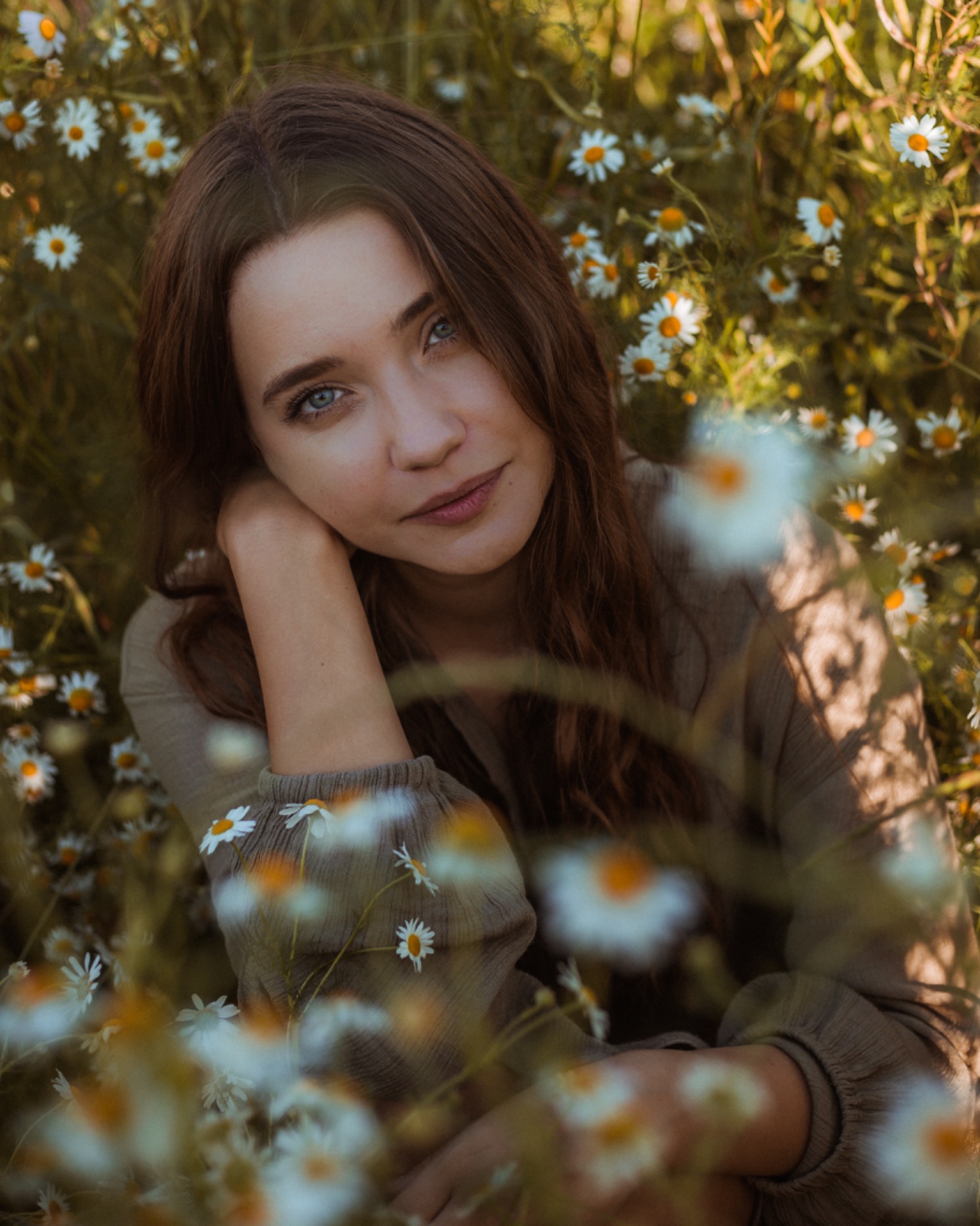 Portret młodej kobiety o niebieskich oczach, otoczonej kwitnącymi margaretkami, opierającej głowę na ręce w naturalnym świetle.