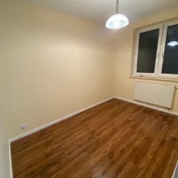 Pusty pokój po remoncie w Bochni z ułożonymi panelami podłogowymi w odcieniach brązu, kremowe ściany, biały grzejnik pod oknem i lampa sufitowa.