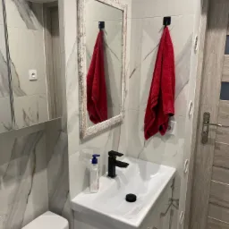 Nowoczesna łazienka z umywalką nablatową, czarną armaturą, lustrem w postarzanej ramie i czerwonym ręcznikiem na tle marmurowych płytek.