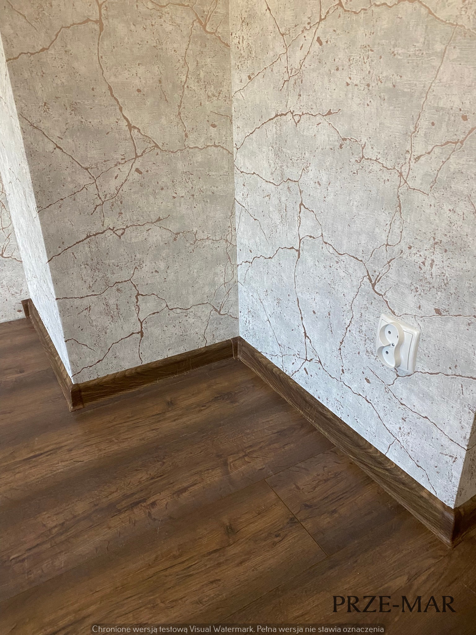 Wykończony róg pokoju z panelami podłogowymi w odcieniach brązu, listwami przypodłogowymi w kolorze paneli i tapetą imitującą popękany beton w odcieniach szarości z brązowymi żyłkami, widoczne...