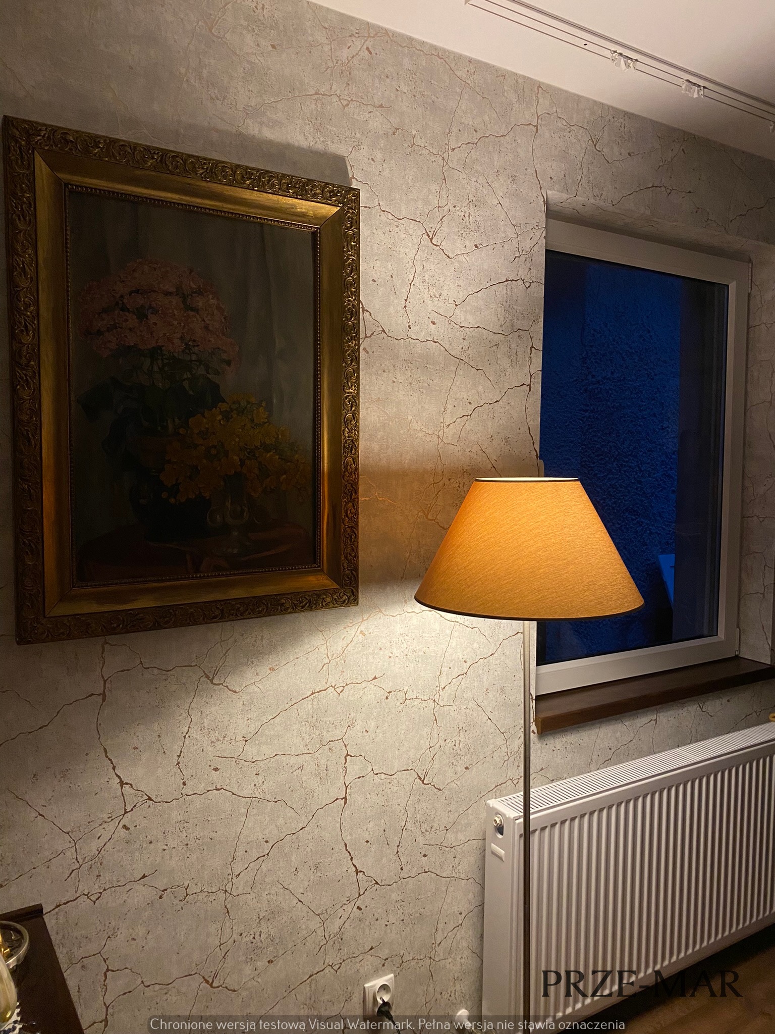 Pokój z tapetą imitującą spękania, oświetlony lampą stojącą z pomarańczowym abażurem, z obrazem w złotej ramie i białym grzejnikiem.
