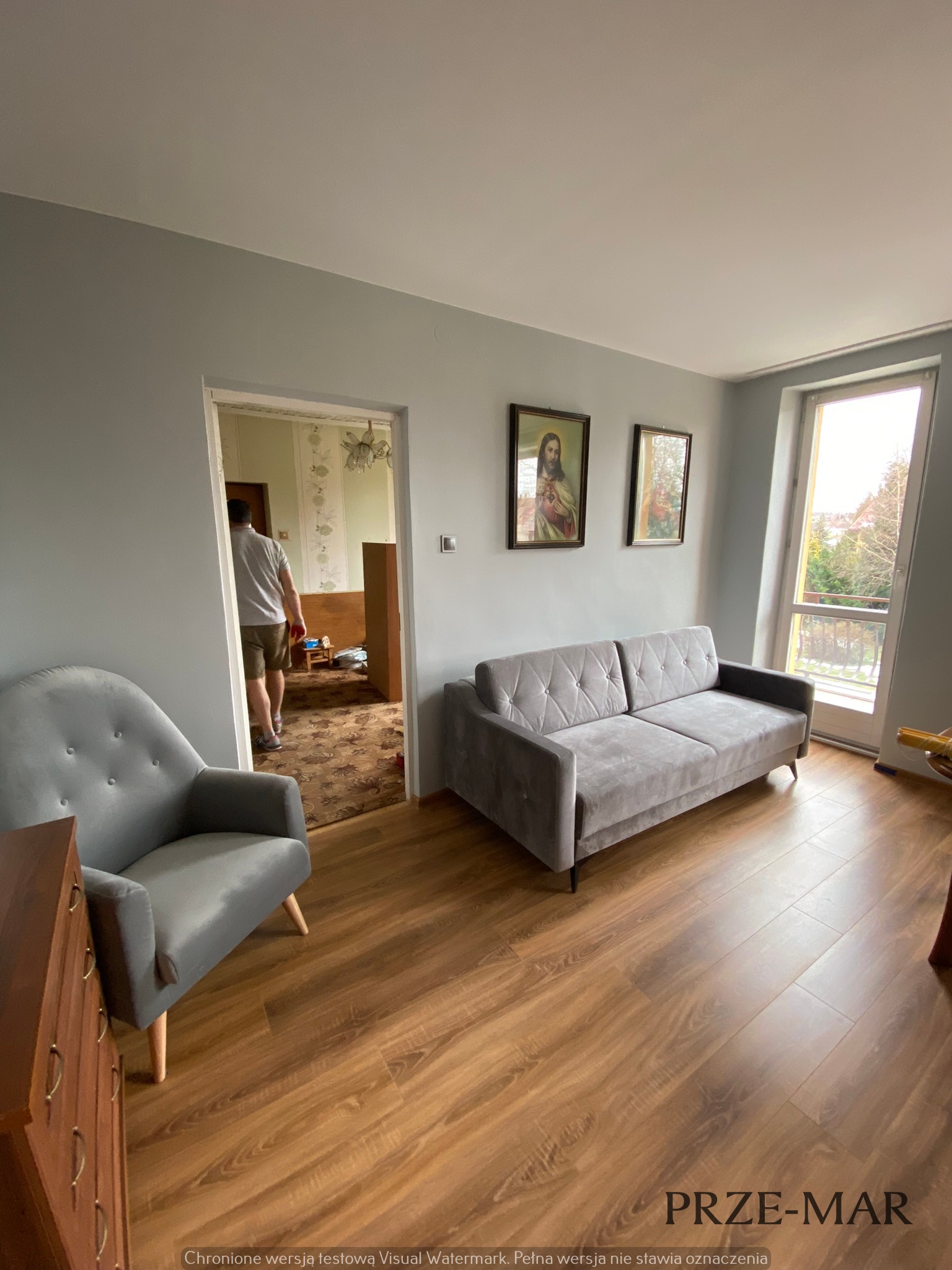 Szare ściany w pokoju z nowo ułożonymi panelami podłogowymi w odcieniach brązu, szara sofa i fotel, widok na pokój obok z wzorzystą tapetą.