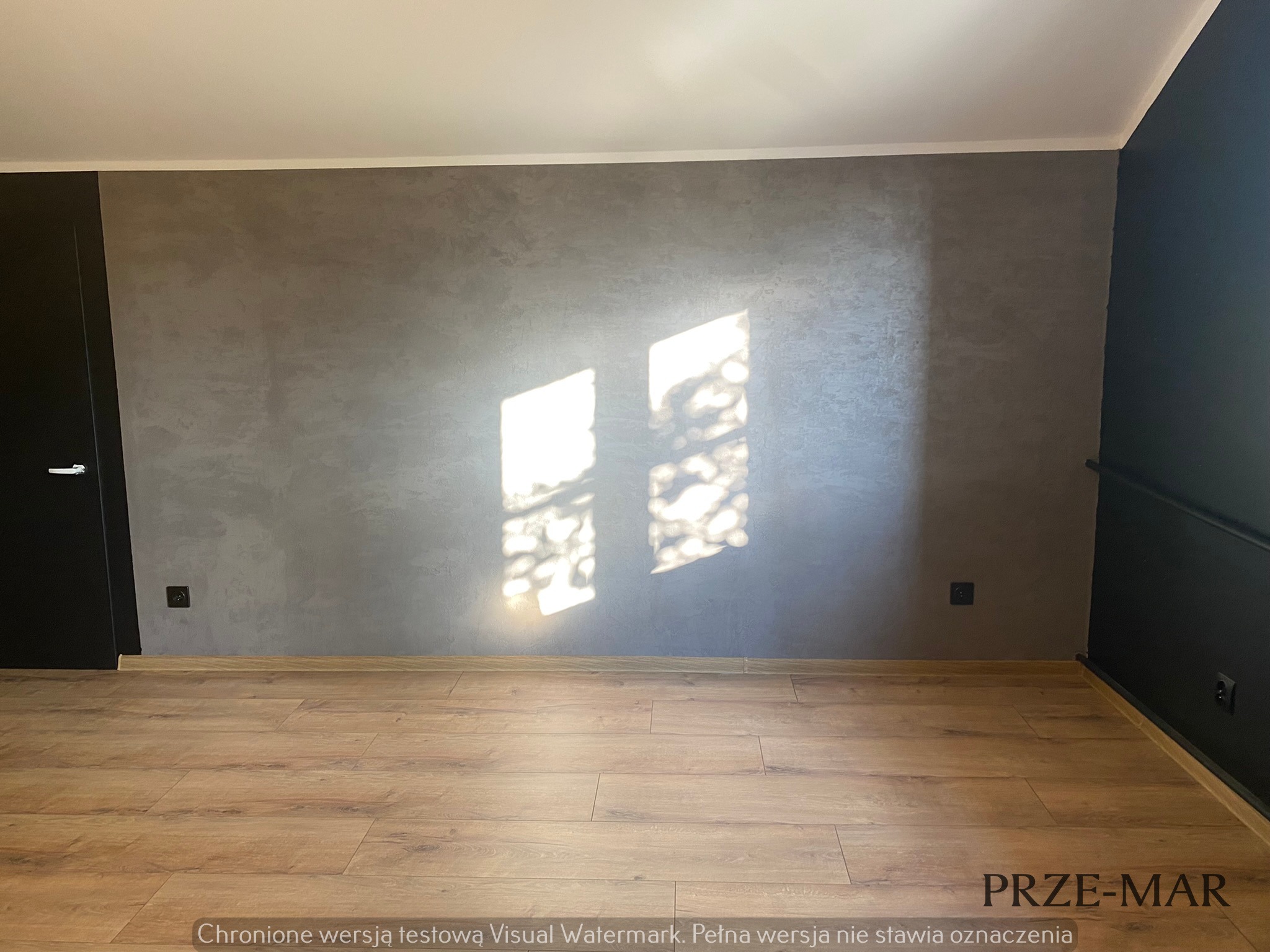 Ściana z efektem dekoracyjnym w odcieniach szarości, oświetlona promieniami słonecznymi wpadającymi przez okno, z widoczną podłogą z paneli imitujących drewno i fragmentem czarnej ściany.