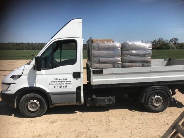 Biały samochód dostawczy z logo firmy ogrodniczej, przewożący palety z ziemią, na tle zielonego pola i lasu.