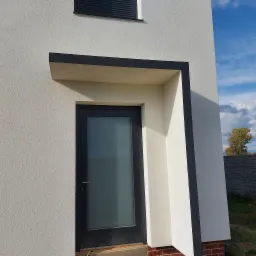 Nowoczesny dom z elewacją w kolorze złamanej bieli, z czarną ramą wokół wejścia i okna, widoczne fragmenty ceglanej podmurówki i błękitne niebo w tle.