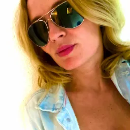 Portret kobiety w okularach typu aviator i dżinsowej kurtce na tle jasnej ściany, ujęcie z dołu.