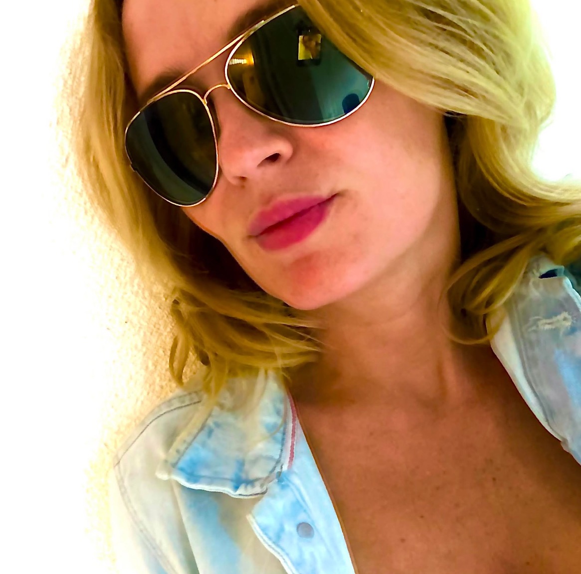 Portret kobiety w okularach typu aviator i dżinsowej kurtce na tle jasnej ściany, ujęcie z dołu.