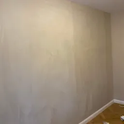 Ściana pokryta dekoracyjną, beżową gładzią w pomieszczeniu z parkietem, widoczna drabina i materiały zabezpieczające podłogę.