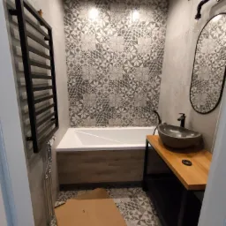 Nowoczesna łazienka z wanną obudowaną drewnem, umywalką w kształcie misy na drewnianym blacie z czarną armaturą i mozaikową ścianą nad wanną, grzejnik drabinkowy po lewej stronie, podłoga w mozaikę...