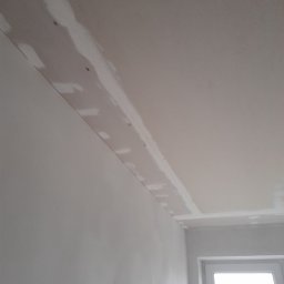 Karol Paziewski - Szpachlowana płyta karton-gips na styku ściany i sufitu, widoczne wkręty i łączenia przed malowaniem, okno w tle.