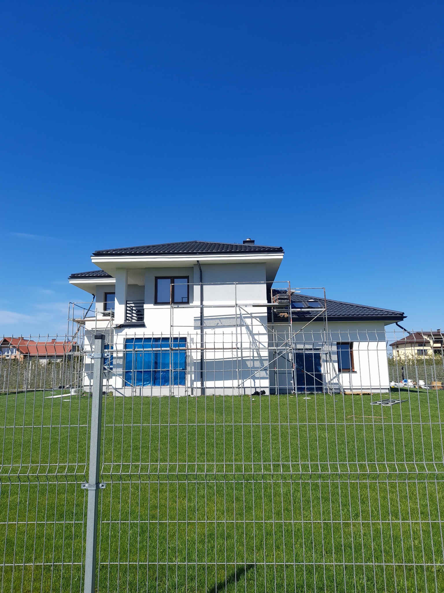 Dom jednorodzinny w Garwolinie w trakcie prac wykończeniowych, widoczne rusztowania i zabezpieczone okna folią, sfotografowany zza ogrodzenia.