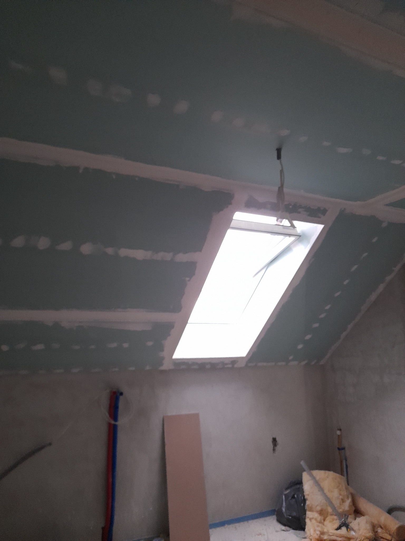 Poddasze w trakcie prac wykończeniowych: surowa płyta karton-gips na skosach i suficie, widoczne łączenia szpachlowane, okno dachowe wpuszczone w konstrukcję, fragment instalacji hydraulicznej...