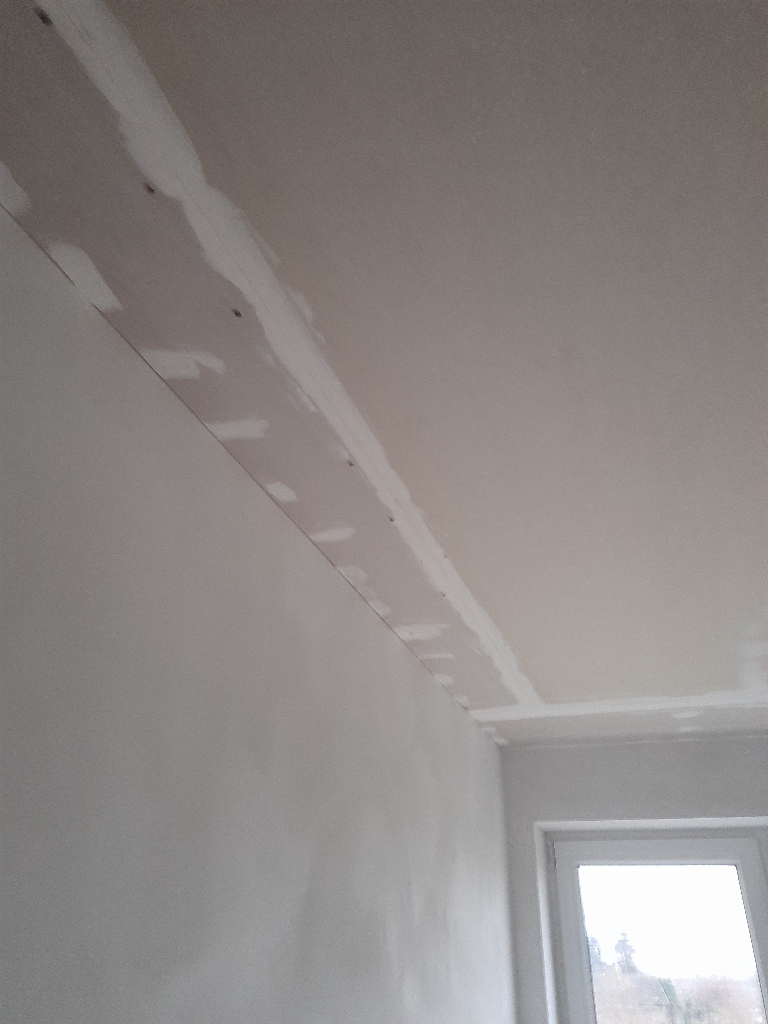 Szpachlowana płyta karton-gips na styku ściany i sufitu, widoczne wkręty i łączenia przed malowaniem, okno w tle.