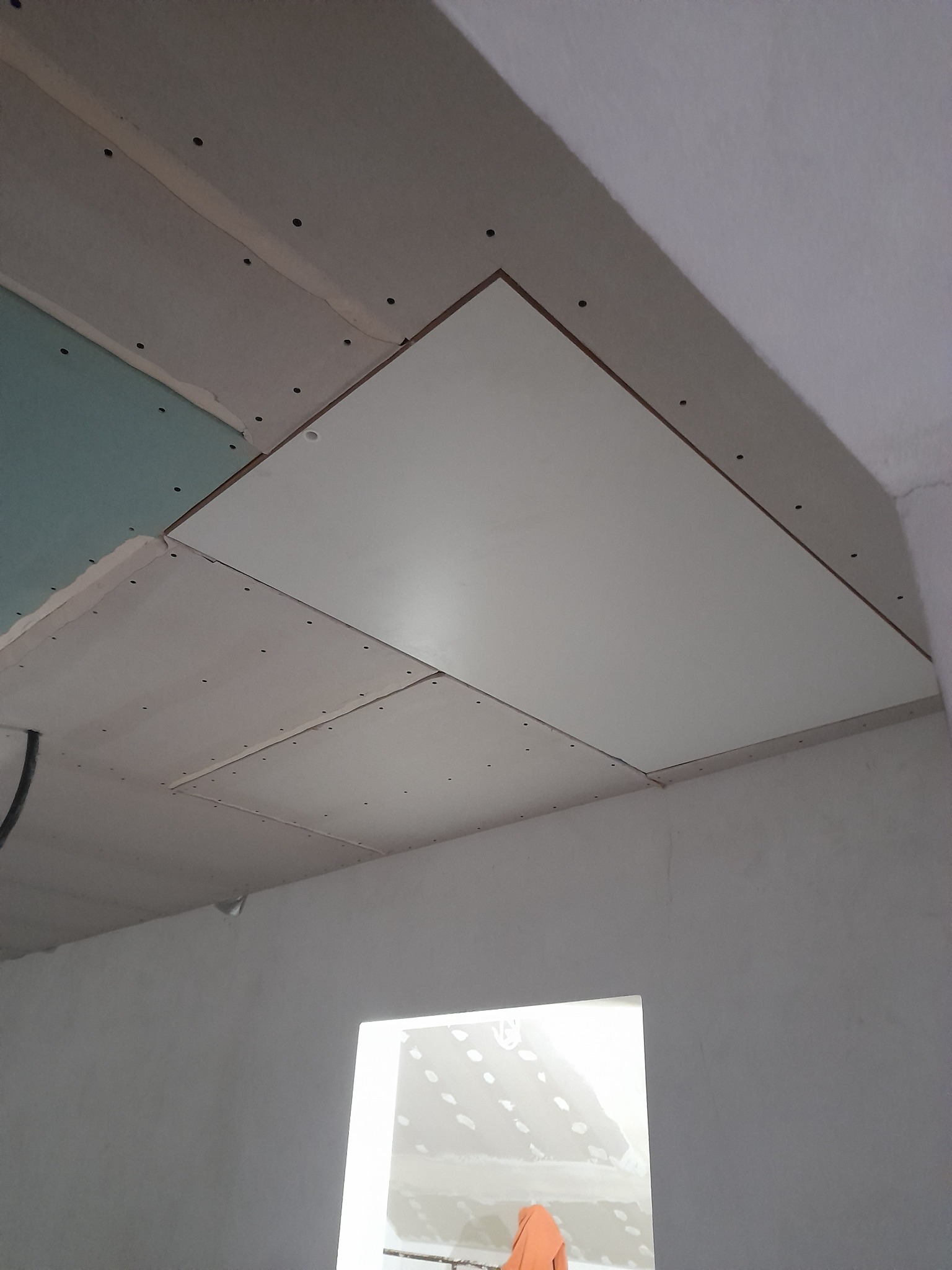 Niedokończony sufit z płyt kartonowo-gipsowych, widoczne łączenia i wkręty, fragment zielonej płyty oraz otwór okienny w tle.
