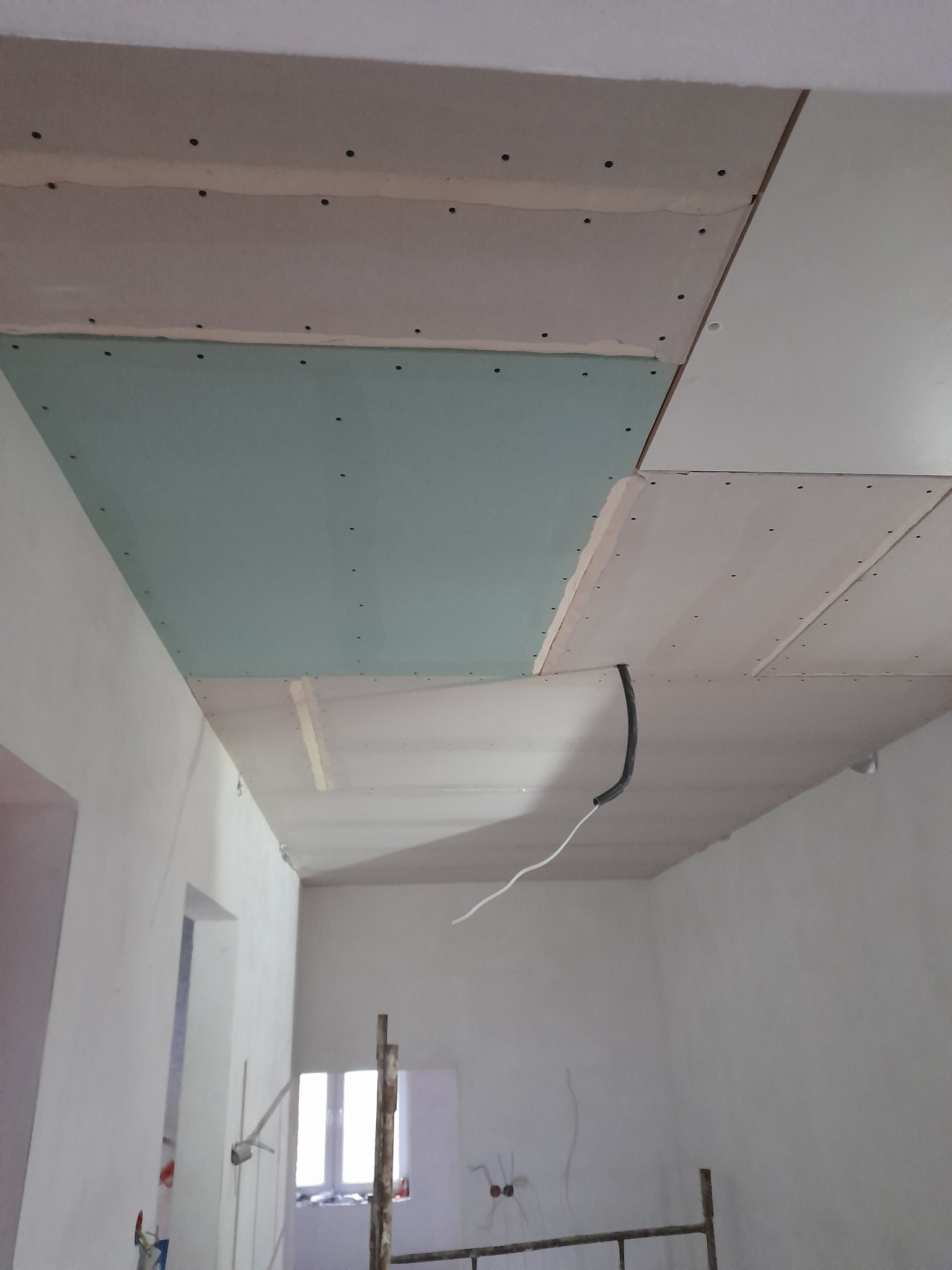 Nierówna powierzchnia sufitu z zamontowanymi płytami kartonowo-gipsowymi, widoczne łączenia i ślady po wkrętach, wystające przewody elektryczne, w tle fragment rusztowania i okno.