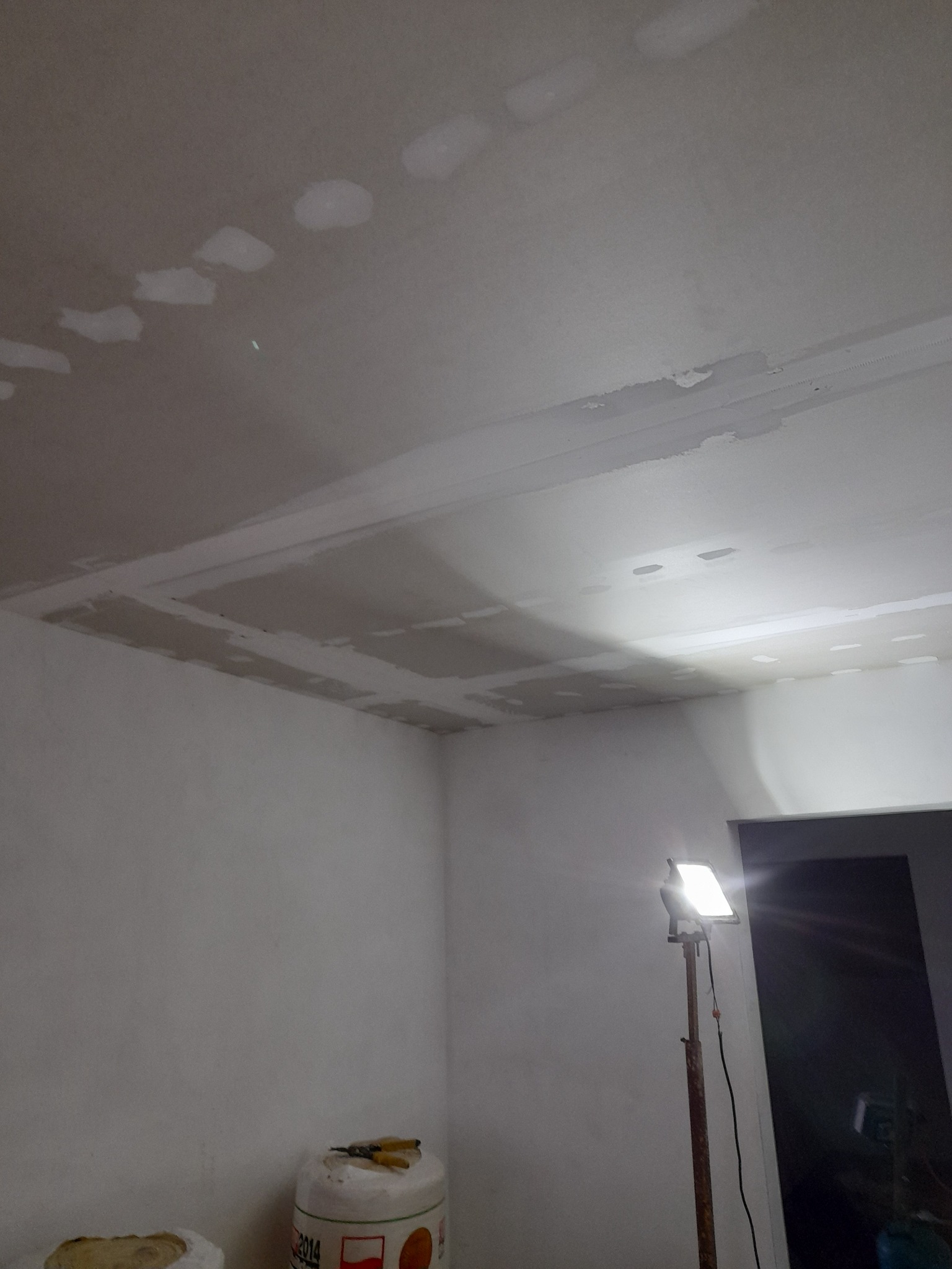 Niedokończony sufit z płyt kartonowo-gipsowych, widoczne łączenia szpachlowane, oświetlone lampą budowlaną na statywie, w tle wiadro z zaprawą i narzędzia.