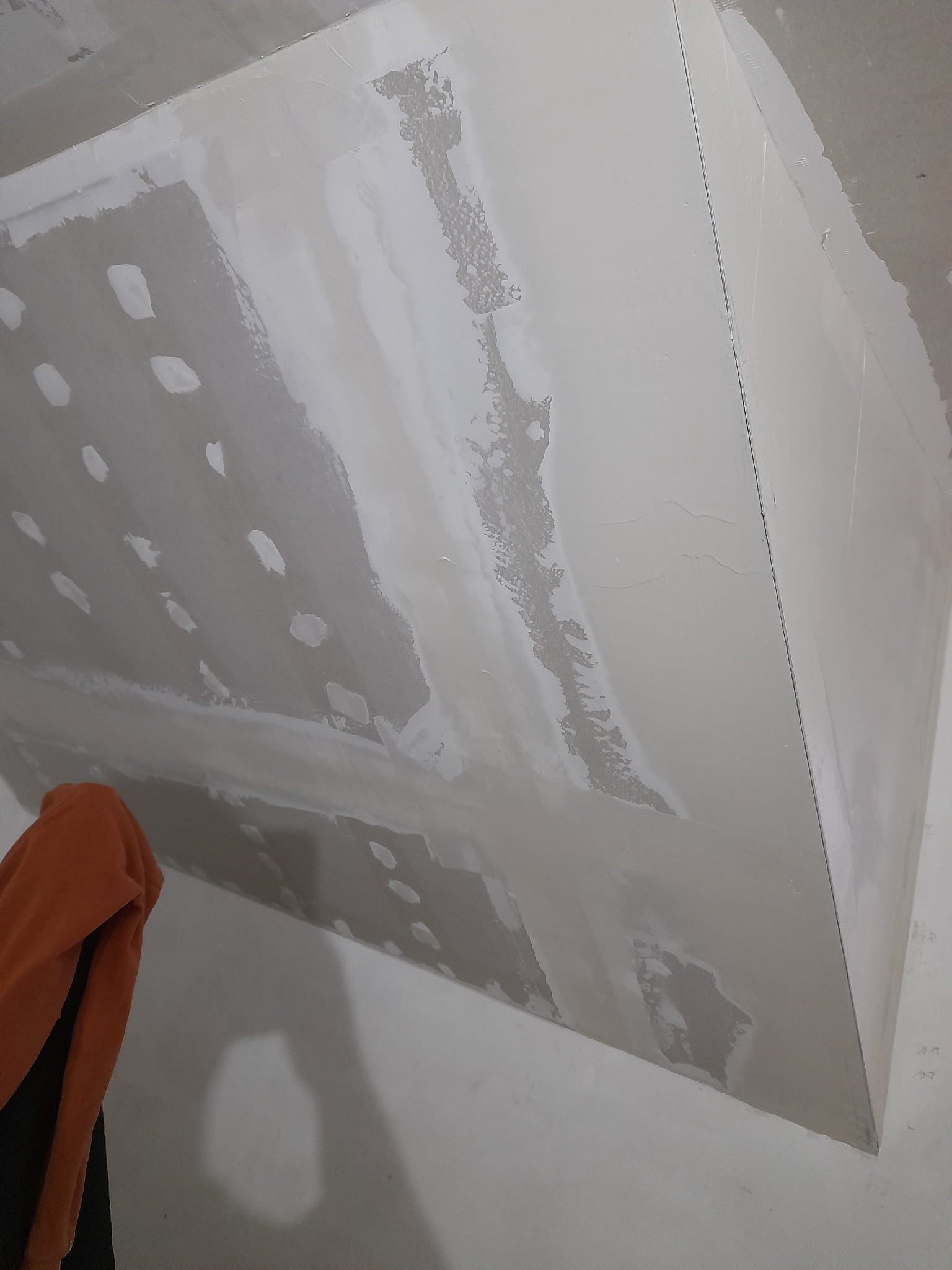 Niedokończona zabudowa z płyty karton-gips na suficie, widoczne łączenia i miejsca po szpachlowaniu, fragment pomarańczowej bluzy w lewym dolnym rogu.