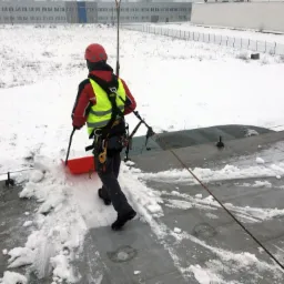 Pracownik w kasku i kamizelce odblaskowej odśnieża płaski dach budynku przemysłowego za pomocą czerwonej łopaty, zabezpieczony linami alpinistycznymi.