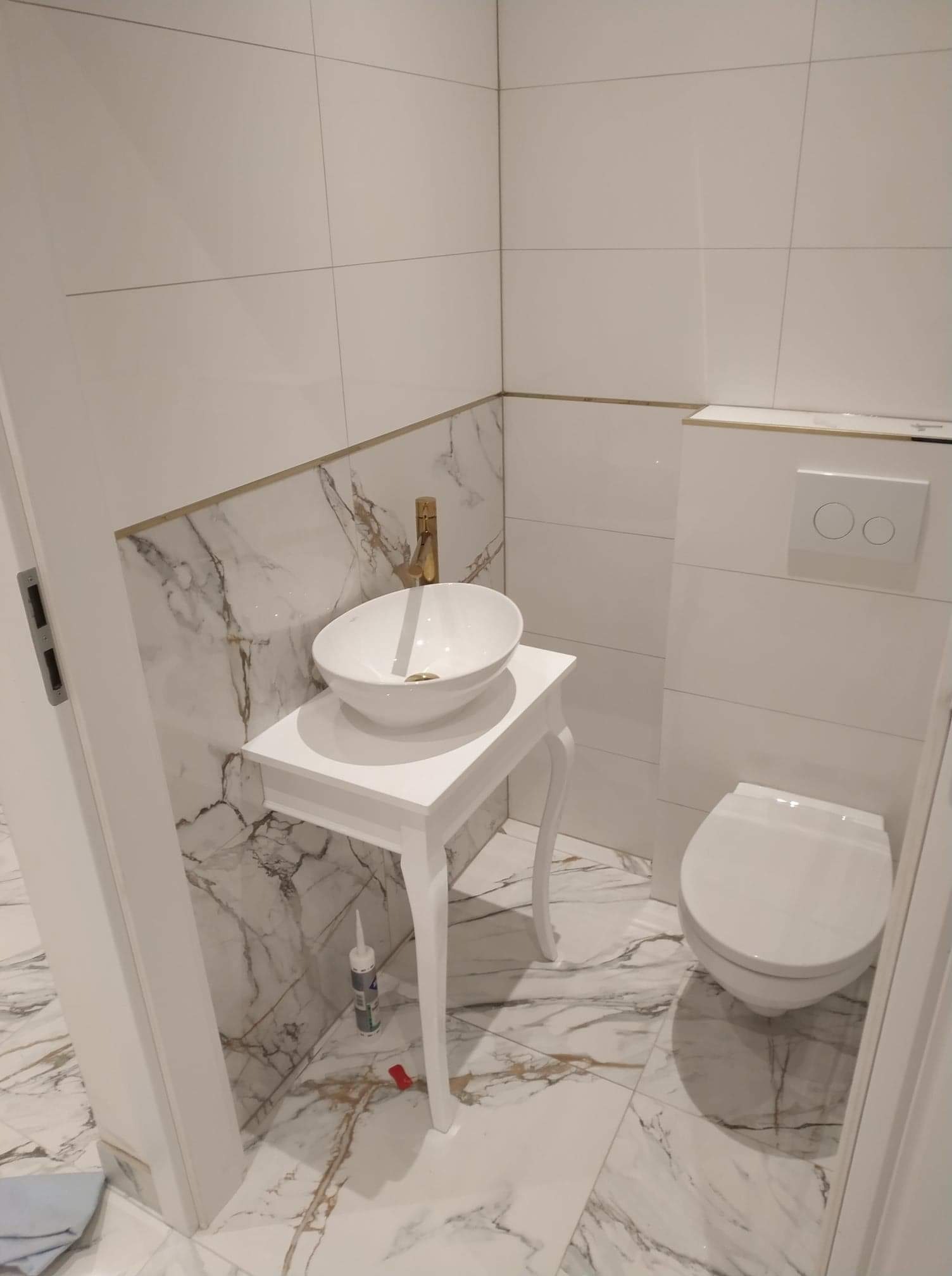 Nowoczesna łazienka z marmurowymi płytkami, białą umywalką na ozdobnym stoliku i złotą baterią. Widoczny fragment toalety i białe płytki na ścianach. Pod stolikiem leży tuba silikonu i czerwony...