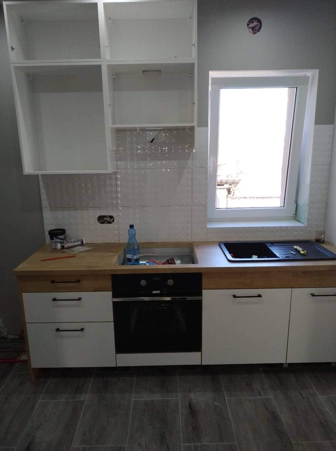 Fragment kuchni w trakcie montażu: białe szafki z drewnianym blatem, czarny piekarnik i zlewozmywak, białe płytki na ścianie oraz widoczne narzędzia i materiały budowlane.