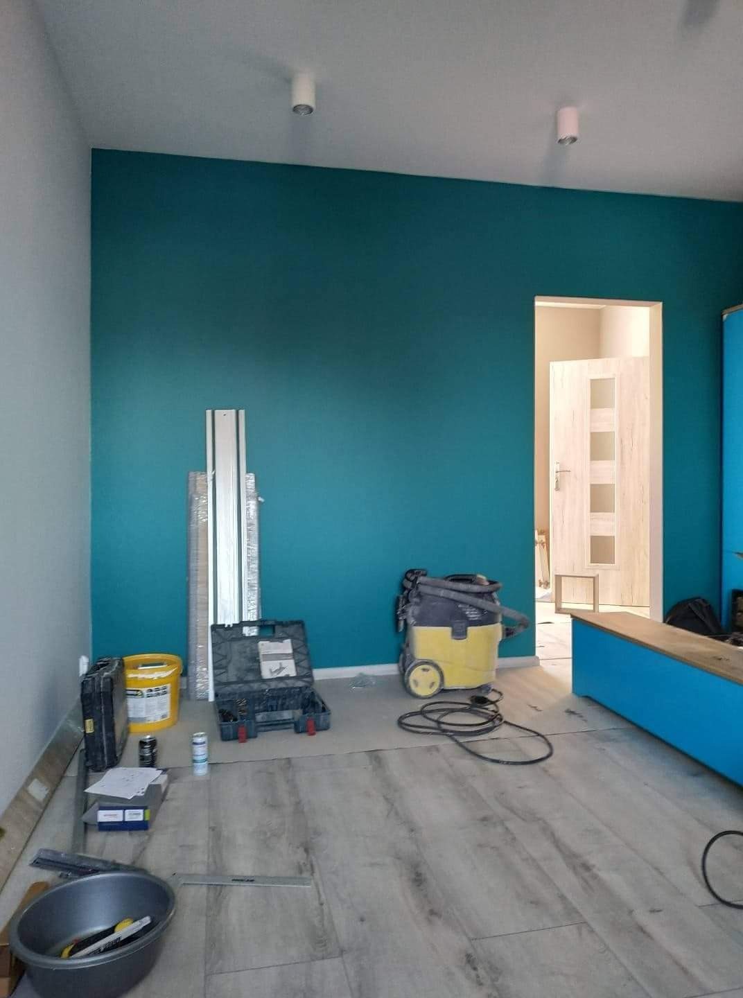 Pokój w trakcie remontu: ściana pomalowana na turkusowo, podłoga z paneli, widoczne narzędzia, wiadro z farbą, odkurzacz przemysłowy i listwy przypodłogowe przygotowane do montażu.
