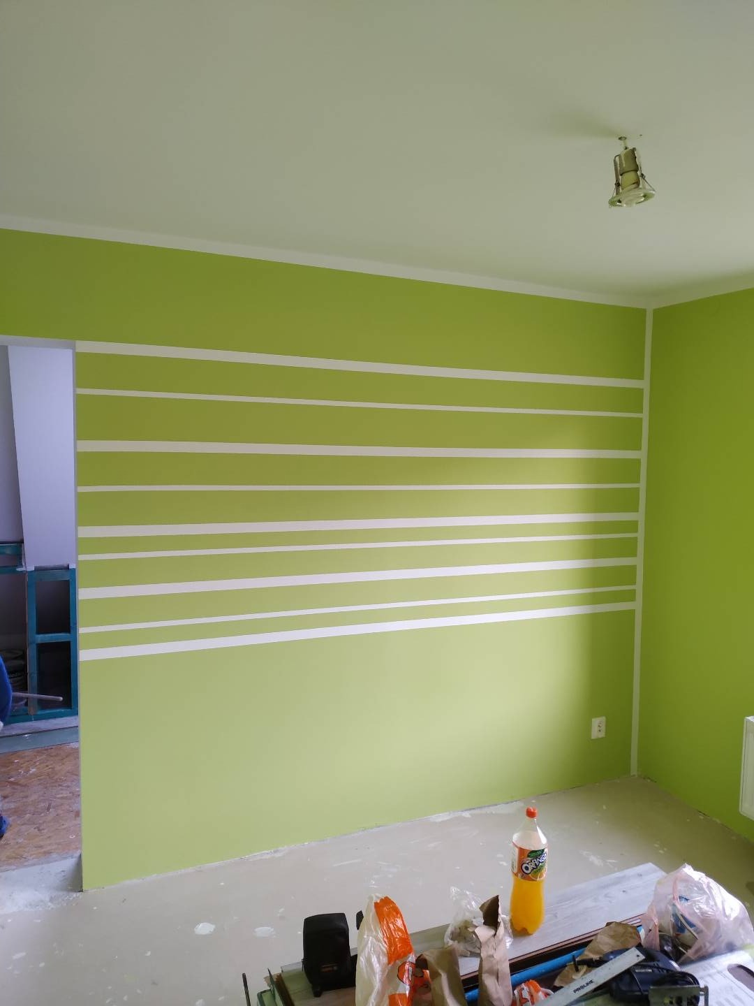 Świeżo pomalowane na jasnozielono ściany z białymi poziomymi pasami w pokoju, widoczne narzędzia malarskie i butelka napoju na podłodze, surowy stan wykończenia.