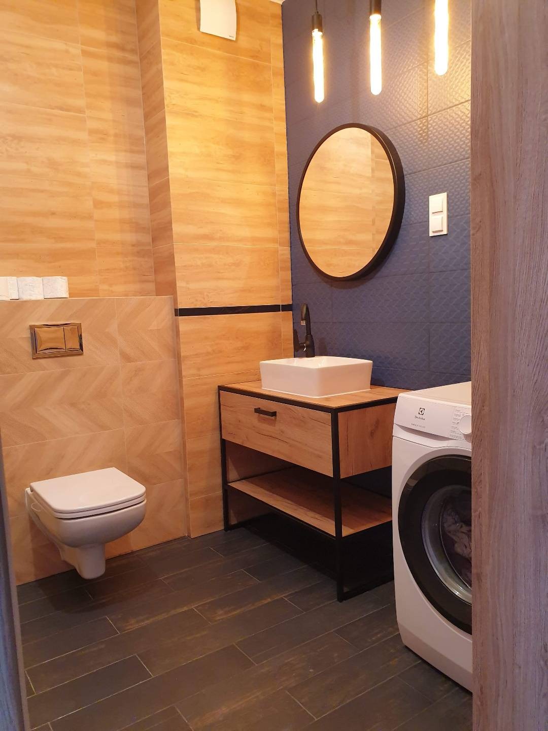 Nowoczesna łazienka z beżowymi płytkami w jodełkę, umywalką na drewnianej szafce z czarnymi metalowymi nogami, okrągłym lustrem i pralką Electrolux.