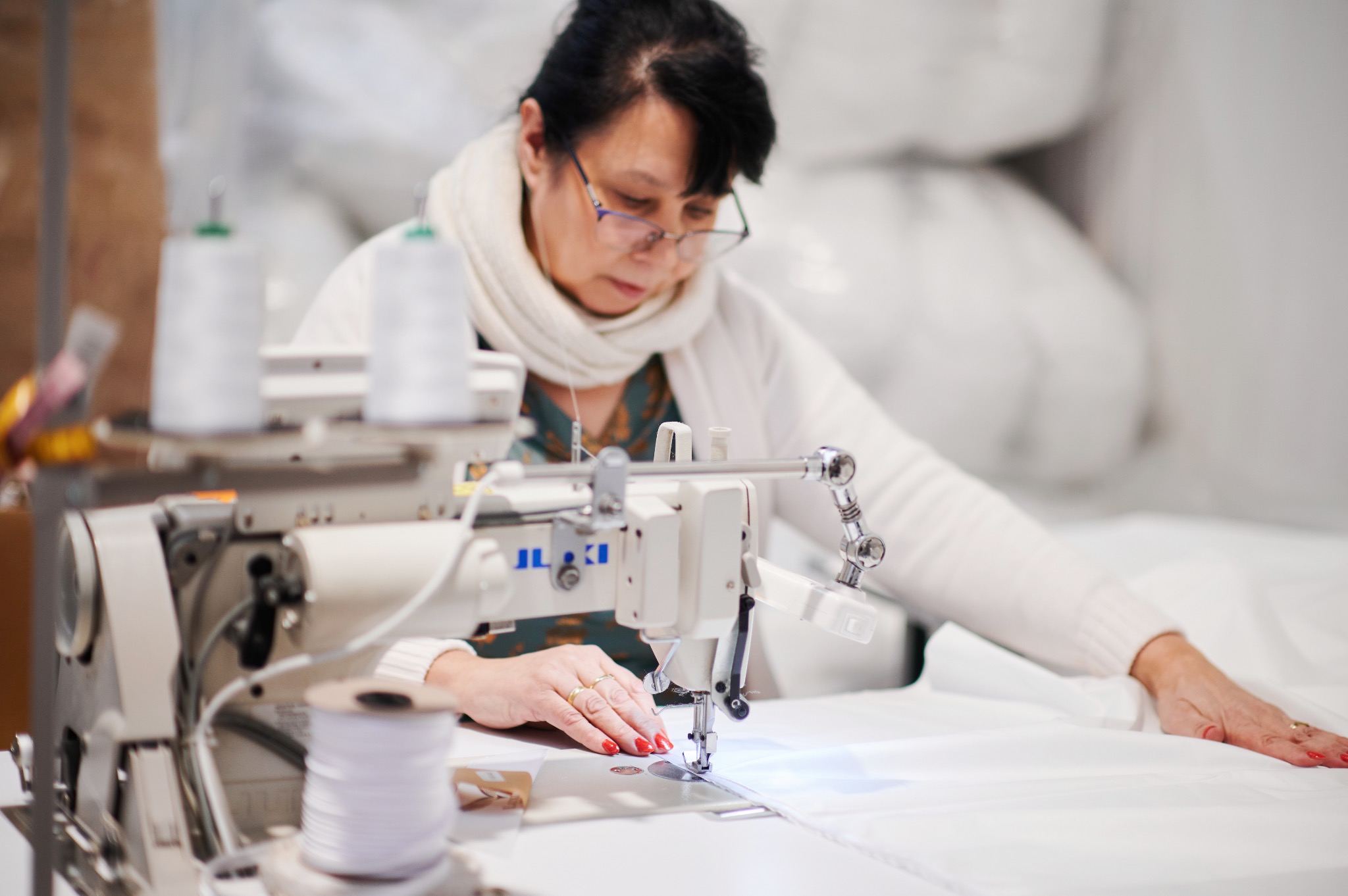 Kobieta w okularach, w trakcie pracy na profesjonalnej maszynie do szycia, szyjąca biały materiał, z widocznymi szpulkami nici i stosem materiałów w tle.