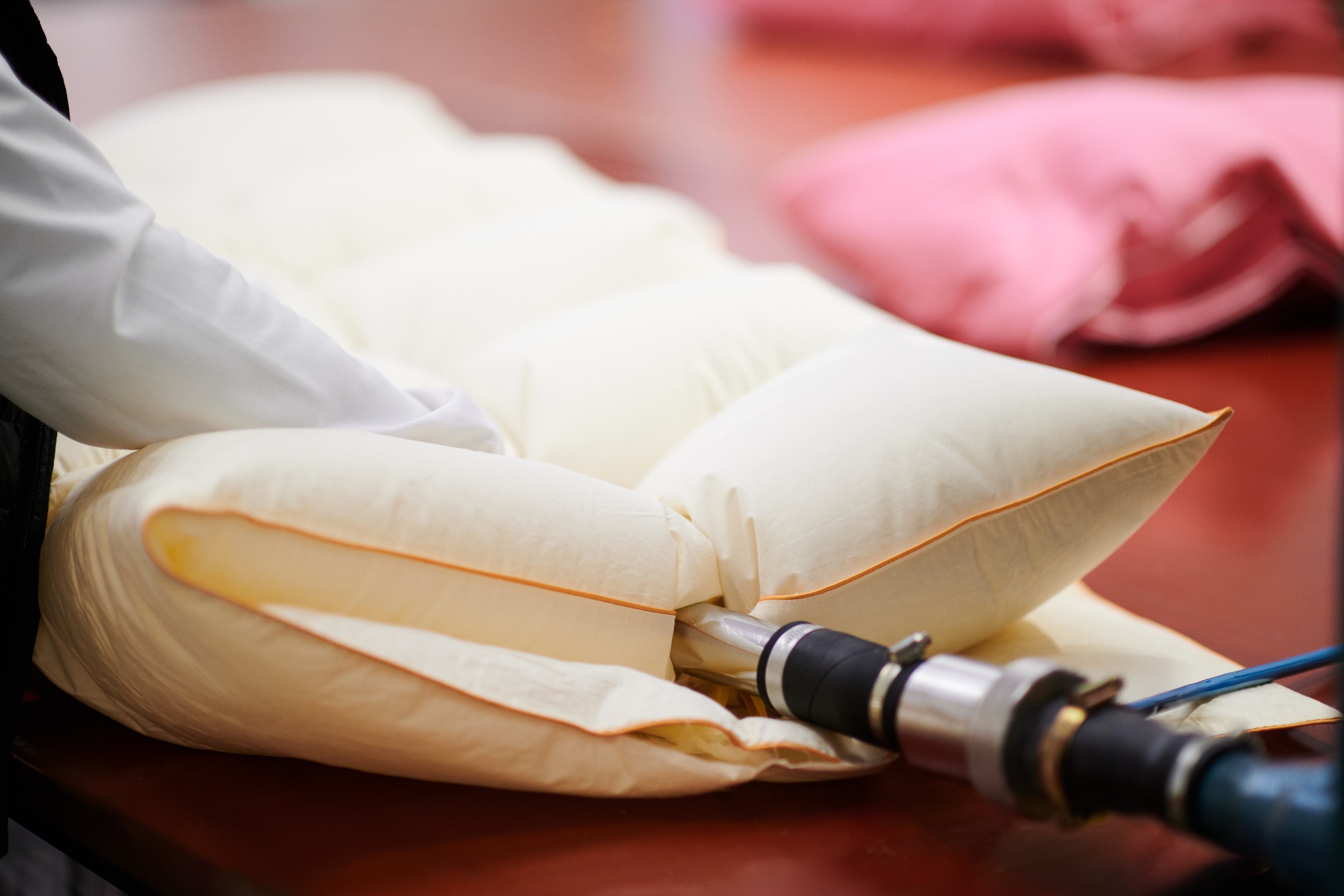 Proces napełniania kremowej poduszki z pomarańczową lamówką za pomocą pneumatycznego urządzenia na drewnianym stole, w tle widoczna różowa poduszka.