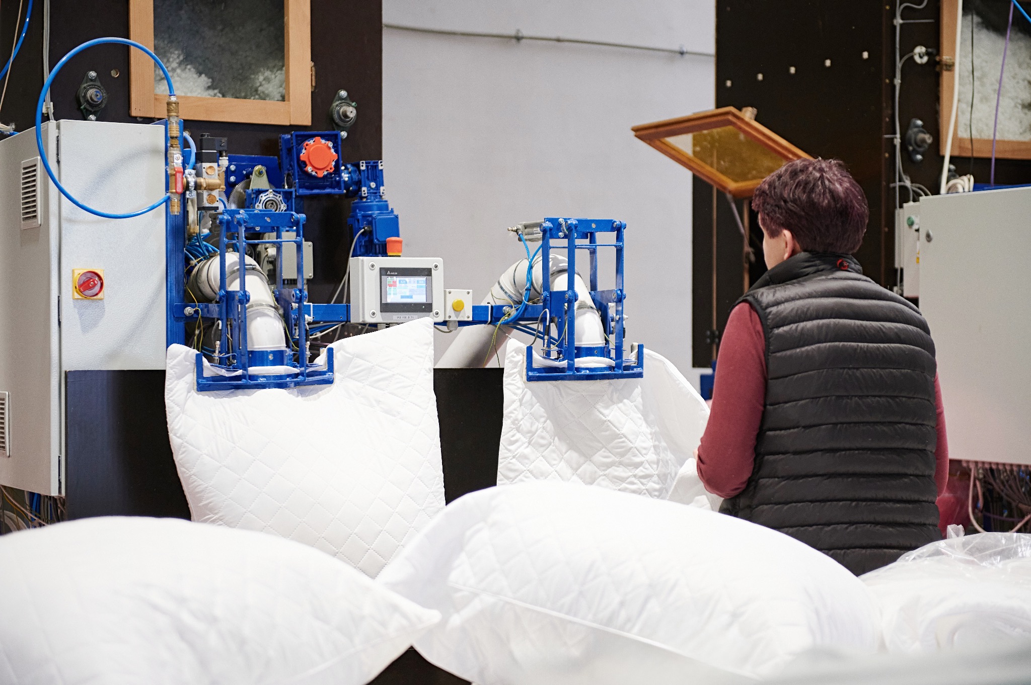 Automatyczna maszyna do napełniania i szycia poduszek, z widocznymi białymi, pikowanymi poduszkami i operatorem w czarnej kamizelce, w tle ciemna ściana z elementami instalacji.
