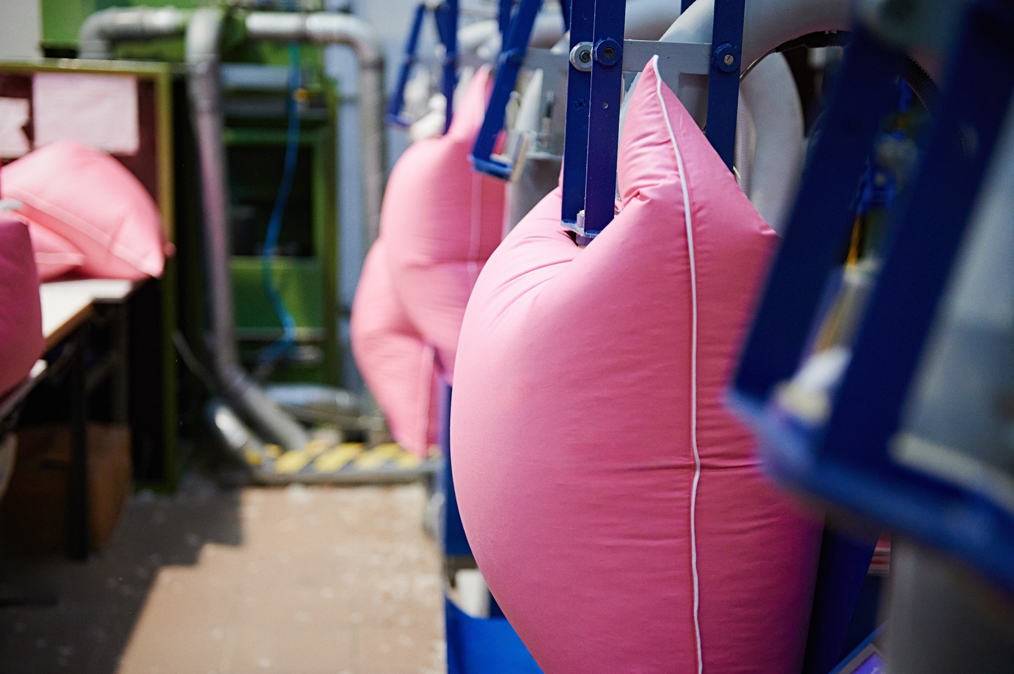 Różowe poduszki z białym obszyciem zawieszone na niebieskim metalowym stelażu w szwalni, widoczne rury i maszyny w tle.