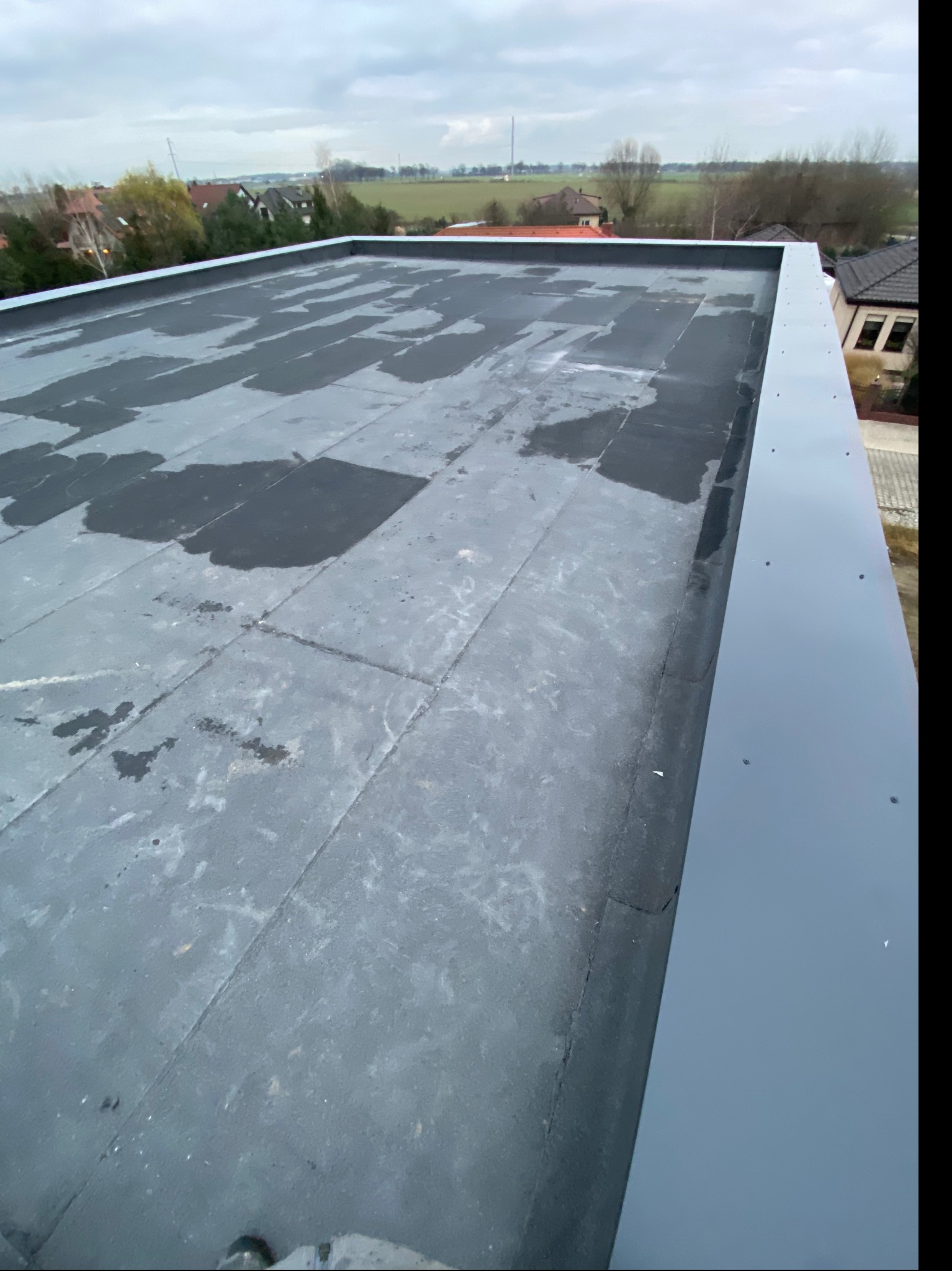 Płaski dach pokryty papą z widocznymi plamami wilgoci i nową, szarą obróbką blacharską na krawędzi, z widokiem na zabudowania i pola w tle, pochmurne niebo.