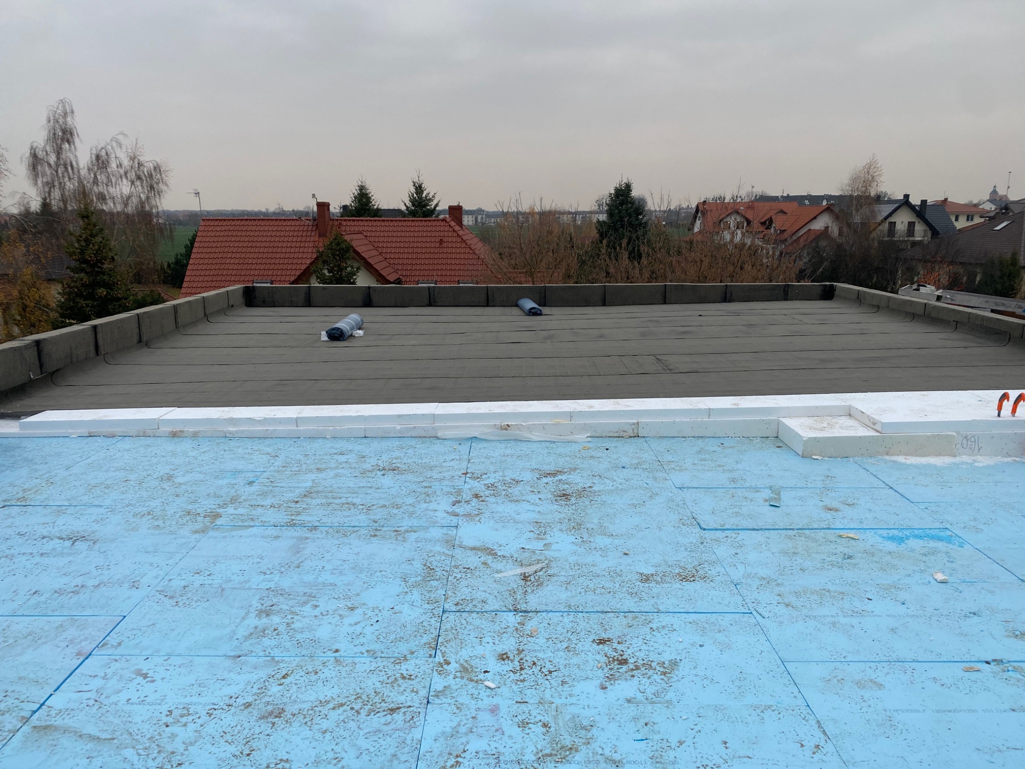 Płaski dach w trakcie izolacji termicznej: widoczna warstwa hydroizolacyjna, białe płyty styropianowe na krawędziach i niebieskie płyty izolacyjne na powierzchni, w tle zabudowa jednorodzinna...