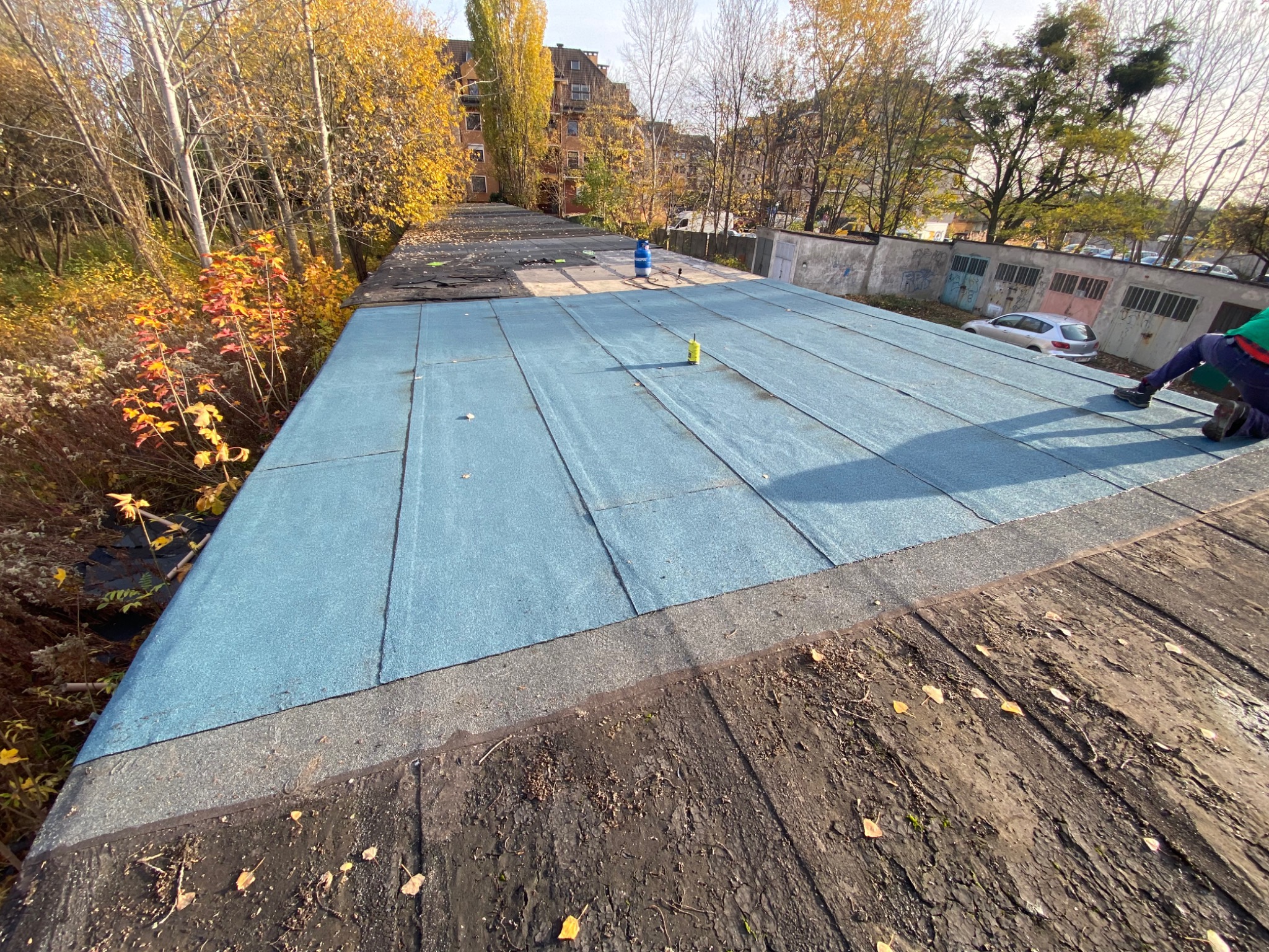 Niebieska izolacja termiczna na płaskim dachu z widocznym fragmentem starego, drewnianego poszycia, butlą gazową i pracownikiem w tle.