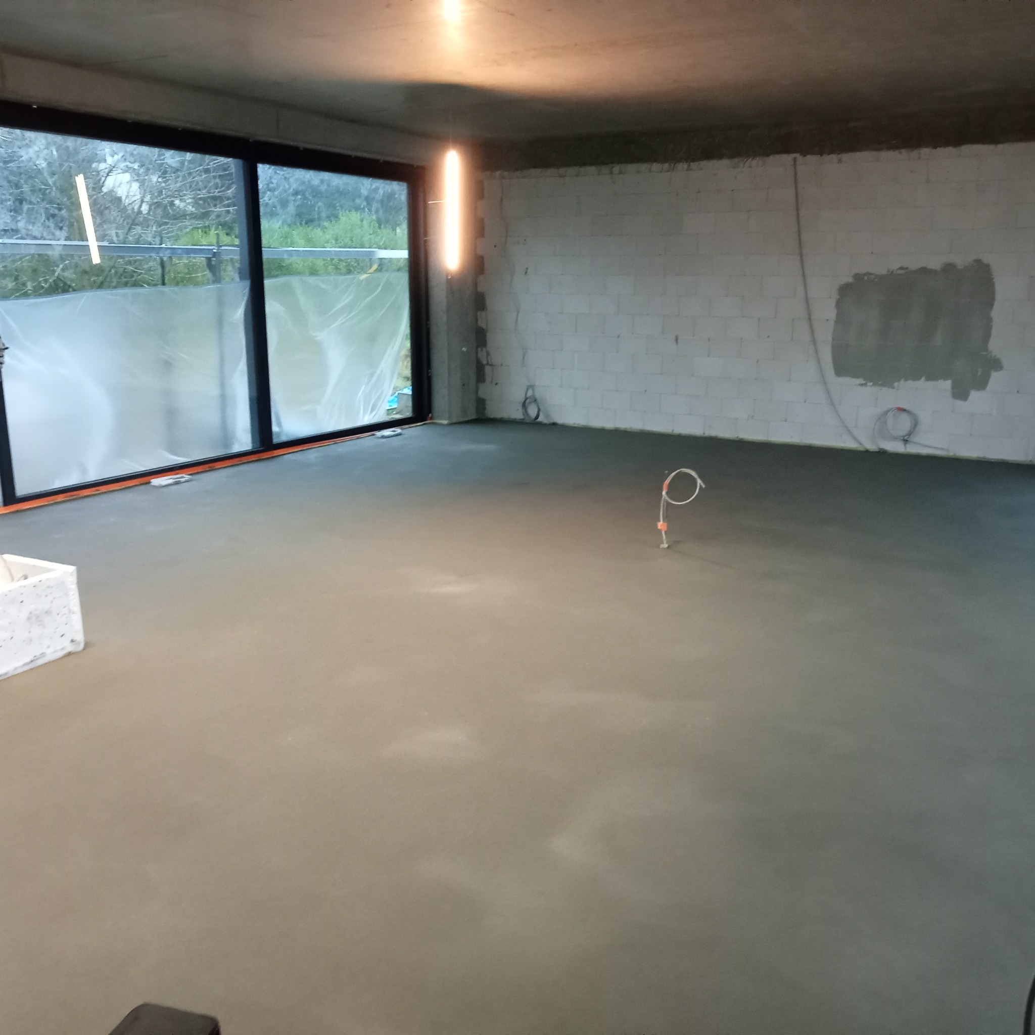 Świeżo wylana, jasnoszara posadzka jastrychowa w surowym wnętrzu z dużym oknem i widocznymi instalacjami elektrycznymi.