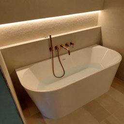 Osoba prywatna - Nowoczesna, wolnostojąca wanna wypełniona wodą, z miedzianą armaturą na tle beżowych płytek i podświetlenia LED. Minimalistyczny design łazienki.