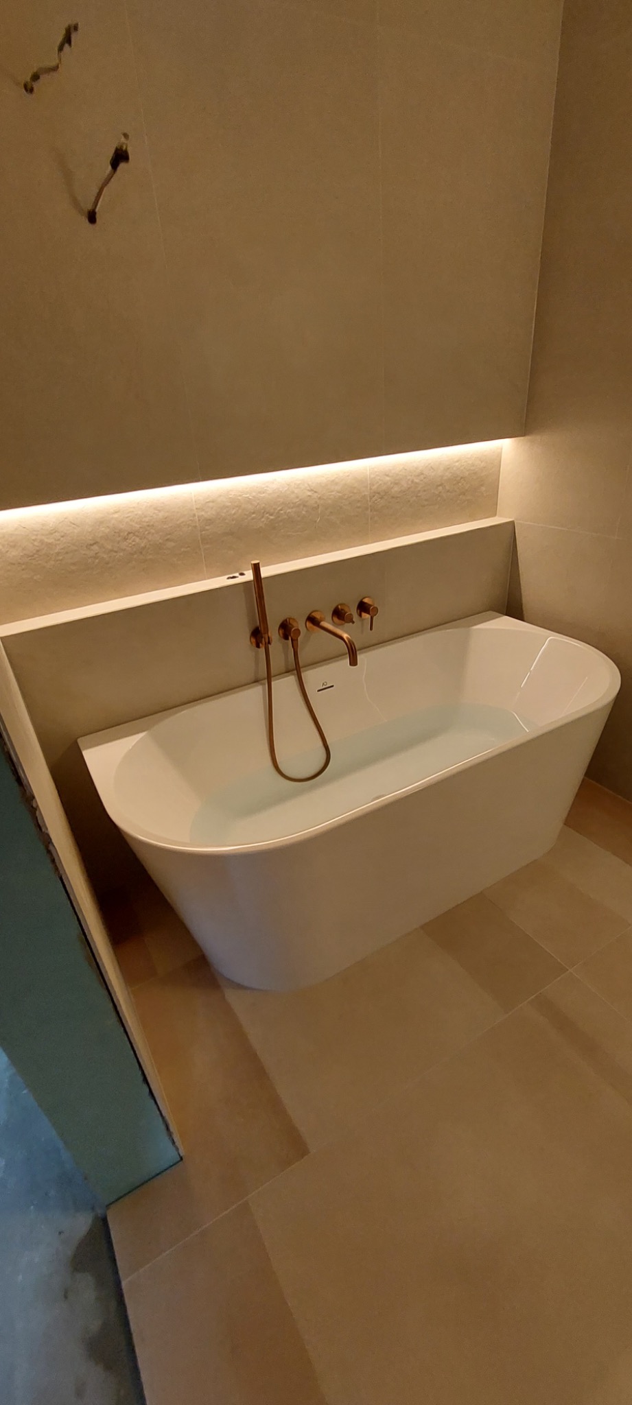 Nowoczesna, wolnostojąca wanna wypełniona wodą, z miedzianą armaturą na tle beżowych płytek i podświetlenia LED. Minimalistyczny design łazienki.