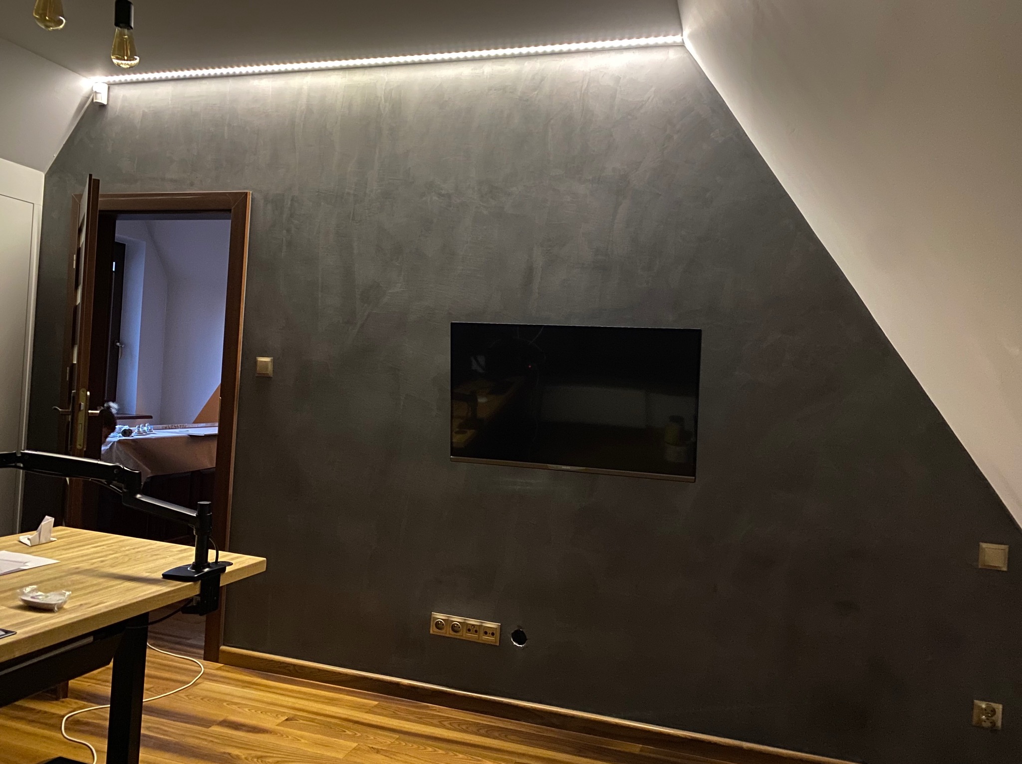 Ściana z efektem dekoracyjnym w odcieniach szarości, podświetlona ledowym paskiem, z zamontowanym telewizorem i widocznymi gniazdkami. W tle otwarte drzwi.