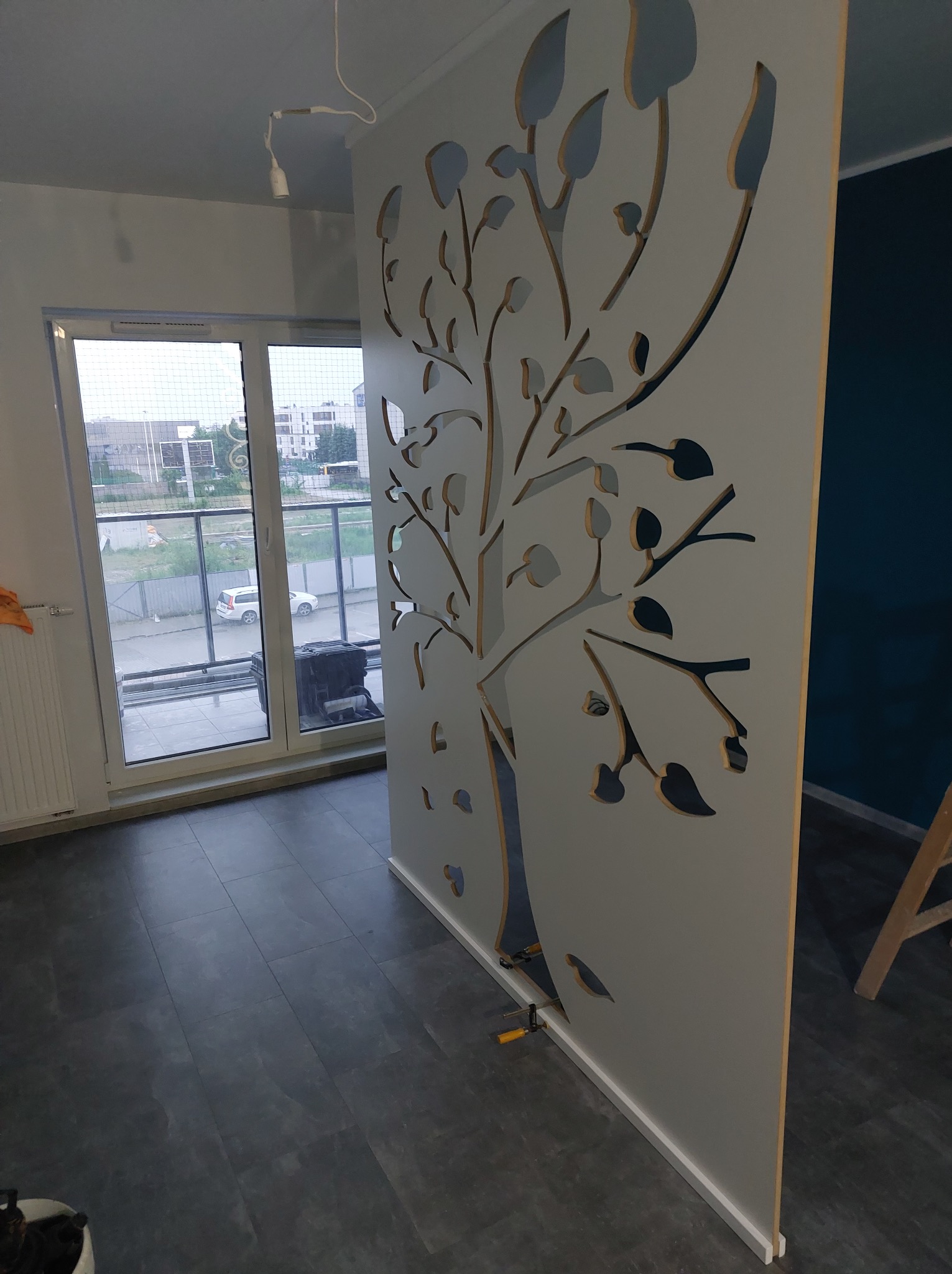Biały ażurowy parawan z motywem drzewa w nowoczesnym wnętrzu. Widoczne narzędzia montażowe, szare panele podłogowe i okno z widokiem na ulicę.