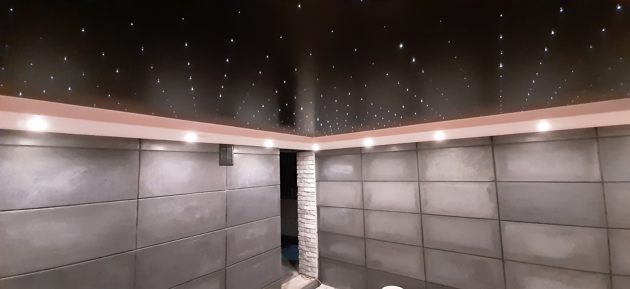 Wykończona łazienka z panelami ściennymi w odcieniach szarości i sufitem imitującym rozgwieżdżone niebo, oświetlonym punktowymi lampami.