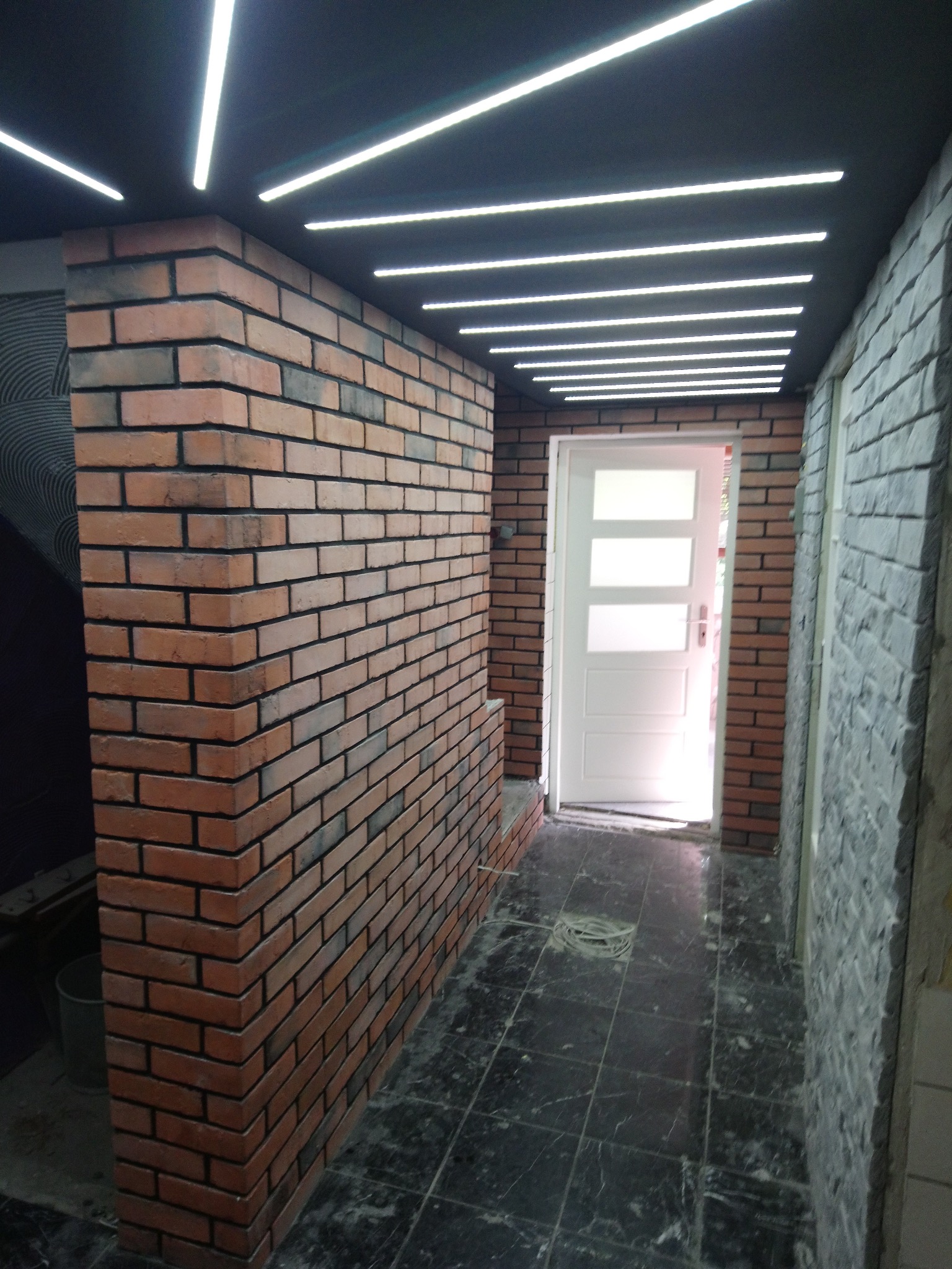 Korytarz z ceglaną ścianą, podświetlanym sufitem i marmurową podłogą. Białe drzwi z szybami w tle. Wykończenie wnętrza w nowoczesnym stylu.