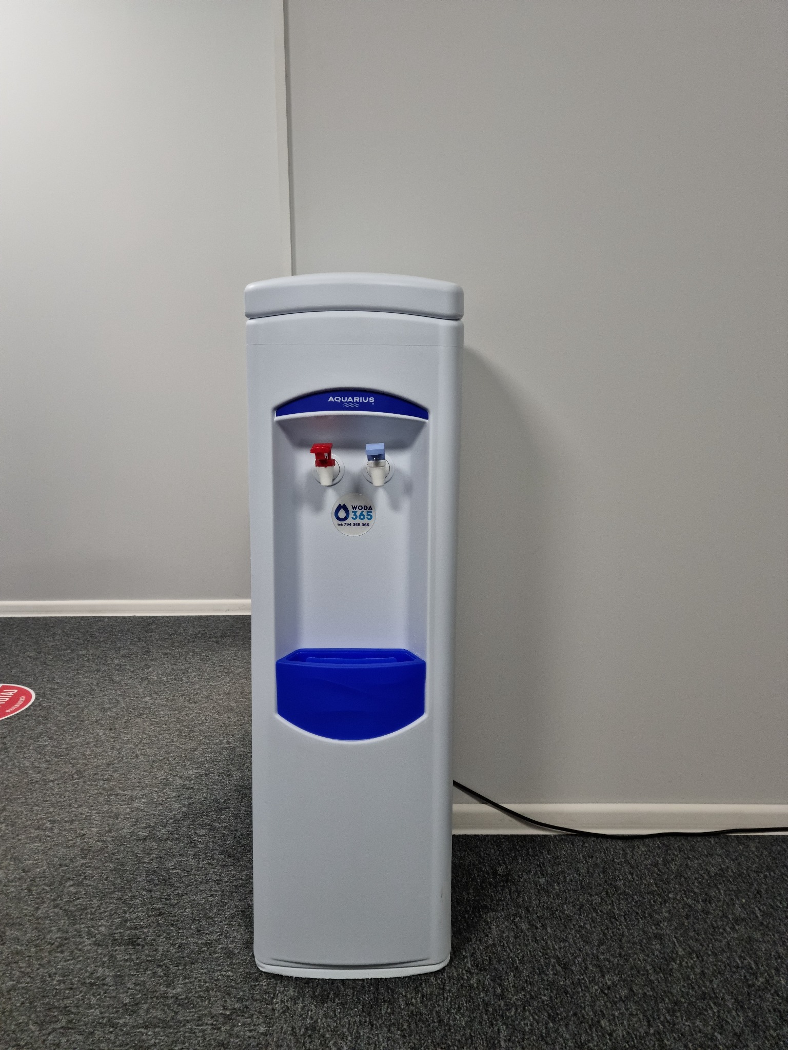 Biały dystrybutor wody Aquarius z niebieskim pojemnikiem na wodę i kranikami ciepłej i zimnej wody, ustawiony na szarym dywanie w biurze.