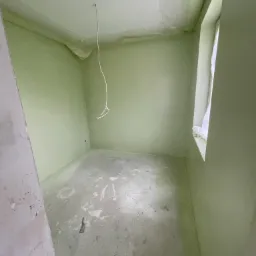 Pokój po malowaniu na jasnozielono, z zaklejonymi folią krawędziami sufitu, wystającym kablem z sufitu i szpachelką leżącą na podłodze.