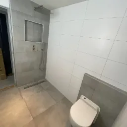 Nowoczesna łazienka z kabiną prysznicową typu walk-in wyłożoną szarymi płytkami i białą ścianą z płytek. W rogu widoczna podwieszana toaleta.