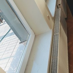 Faktoria Kamienia Grudzień Monika Juda - Białe grzejniki żeberkowe zamontowane pod oknem w jasnym pomieszczeniu z drewnianą podłogą, widoczne ślady czyszczenia okna.