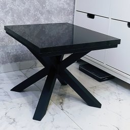 Faktoria Kamienia Grudzień Monika Juda - Czarny stolik kawowy z blatem o fakturze kamienia, z krzyżowymi nogami, ustawiony na marmurowej podłodze, na tle białych szafek i szarej ściany.