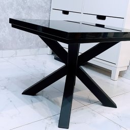 Faktoria Kamienia Grudzień Monika Juda - Czarny stolik kawowy z połyskującym blatem i geometrycznymi nogami na tle marmurowej podłogi i białej komody.