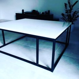 Faktoria Kamienia Grudzień Monika Juda - Nowoczesny stolik kawowy z białym blatem przypominającym konglomerat i czarną, stalową ramą, ustawiony na szarych płytkach podłogowych w minimalistycznym salonie.