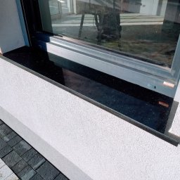 Faktoria Kamienia Grudzień Monika Juda - Czarny, błyszczący parapet zewnętrzny z kamienia, zamontowany pod oknem z szarą ramą, widoczny fragment elewacji budynku z szarą kostką brukową.